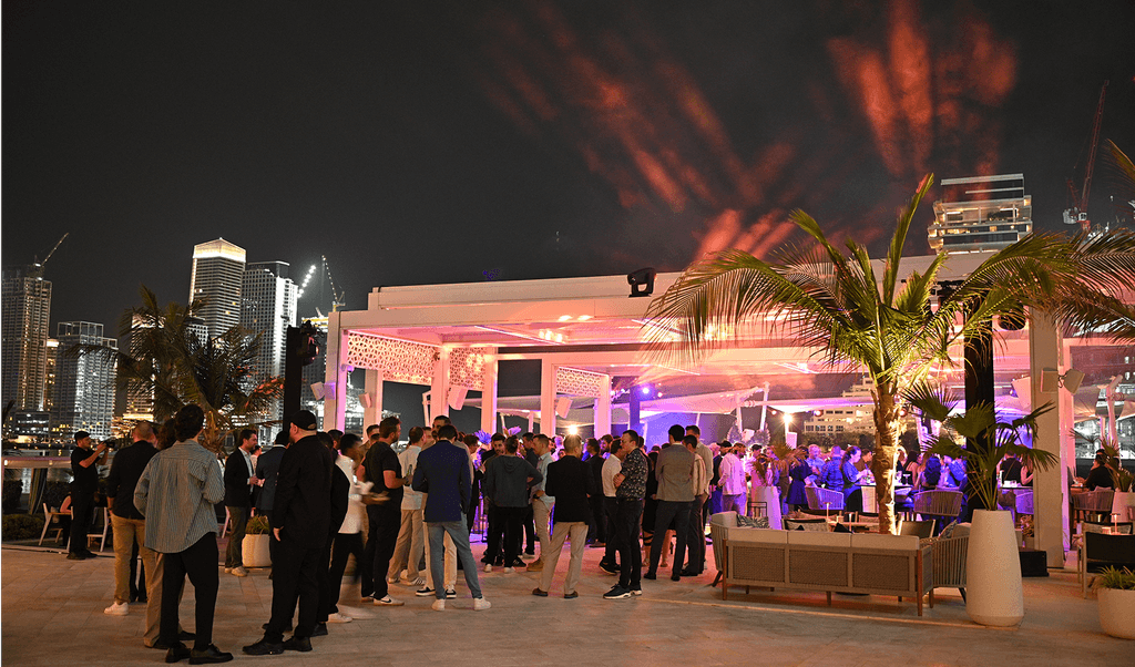 A lively party unfolds at night, with a crowd of people gathered under ambient lighting, surrounded by palm trees, with a city skyline glowing in the background.