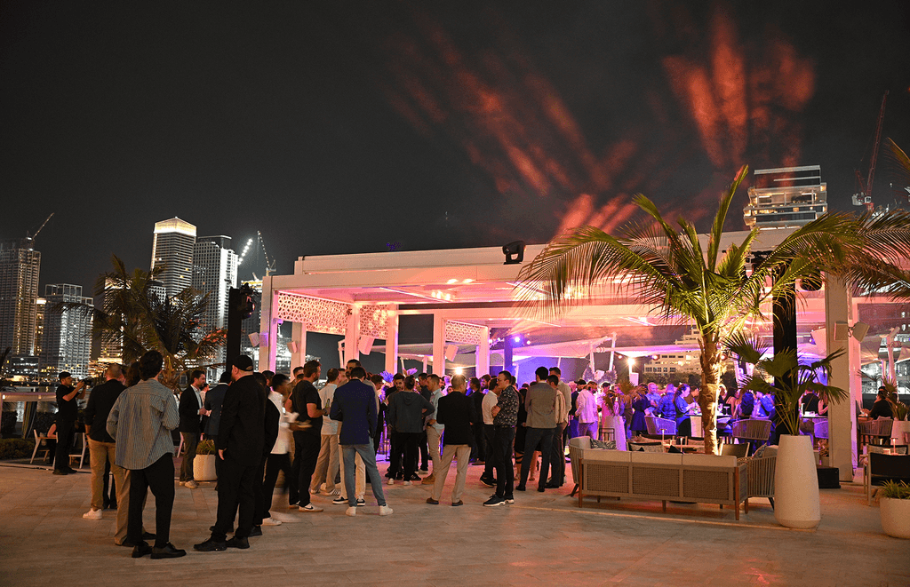 A lively outdoor event in Dubai features elegantly dressed guests mingling under palm trees, with colorful lights and city skyscrapers in the background, as they enjoy dinner and drinks.
